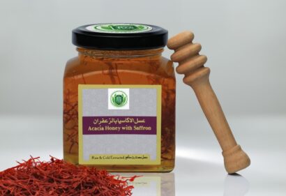 Alt text: A jar of honey with saffron and a wooden stick next to it. The jar is labeled "Acacia Honey with Saffron" and "Raw & Cold Extracted." There are also regions of Simply The Great Food Vegan Acacia Honey and Safron 150g, Simply The Great Food Vegan Acacia Honey and Safron 500g, and Simply The Great Food Vegan Sidr Honey 150g.